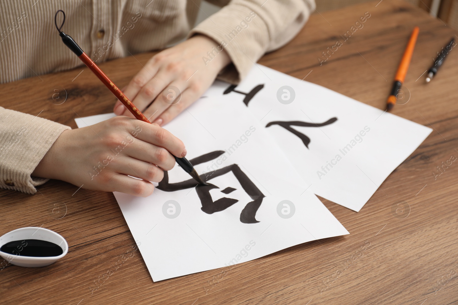 Photo of Calligraphy. Woman with brush and inkwell writing words Bird, River and Entrance in Chinese on paper at wooden table, closeup