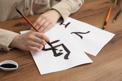 Calligraphy. Woman with brush and inkwell writing words Bird, River and Entrance in Chinese on paper at wooden table, closeup