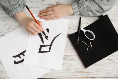 Photo of Calligraphy. Woman with brush and inkwell practicing writing Chinese hieroglyphs on paper at wooden table, top view