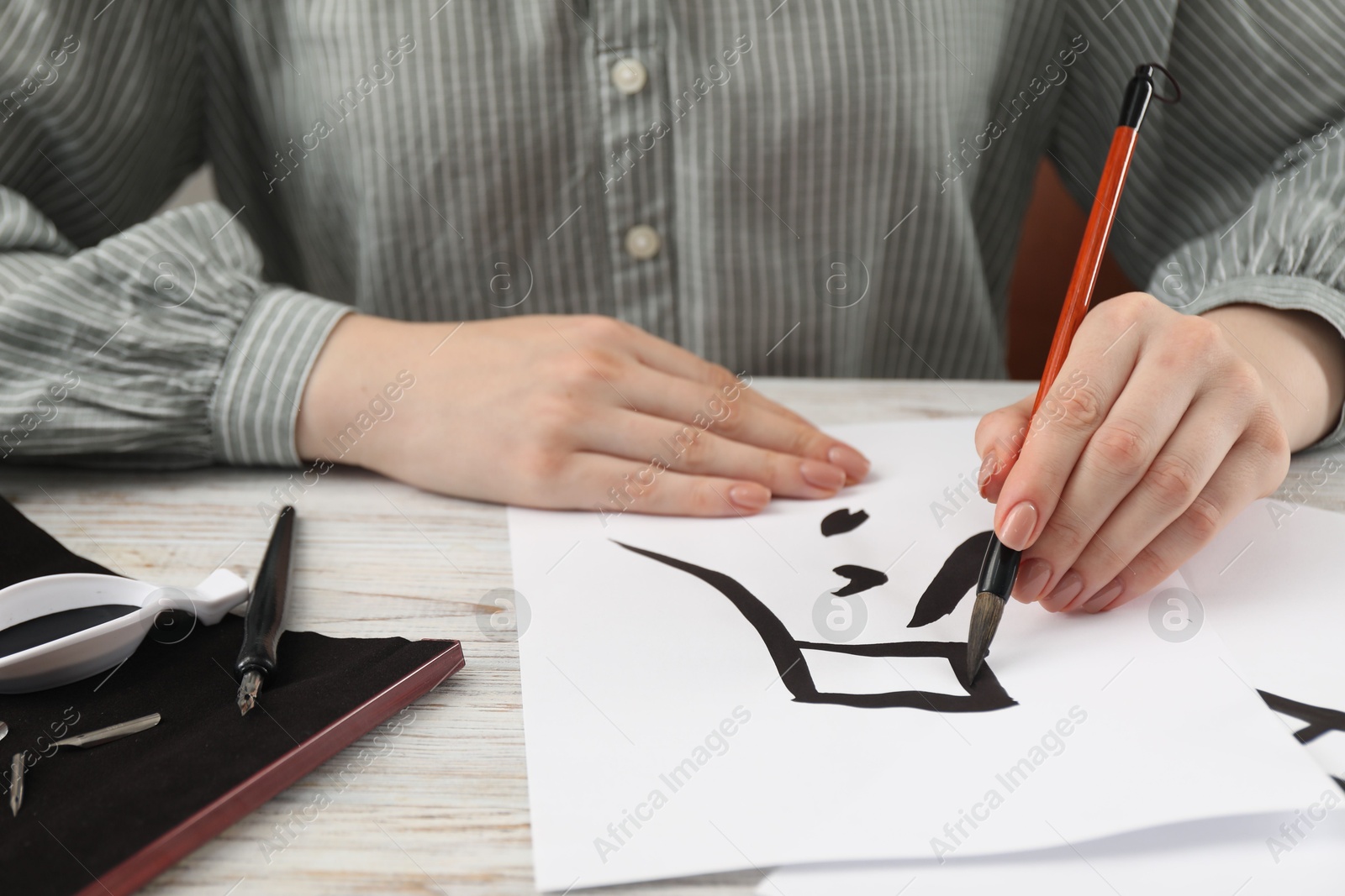 Photo of Calligraphy. Woman with brush and inkwell writing word Finish in Chinese hieroglyphs on paper at white wooden table, closeup