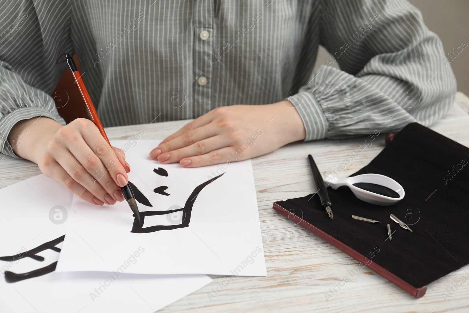 Photo of Calligraphy. Woman with brush and inkwell practicing writing Chinese hieroglyphs on paper at white wooden table, closeup