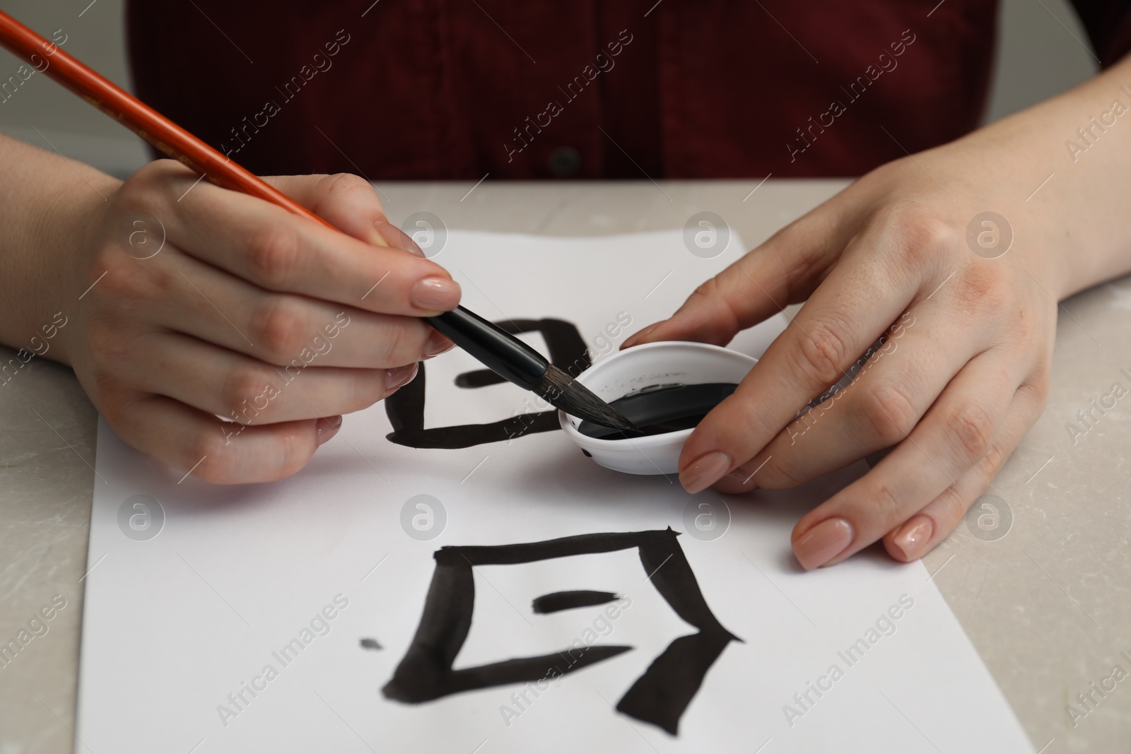 Photo of Calligraphy. Woman dipping brush into inkwell at light marble table, closeup. Words Speak and Day in Chinese on paper