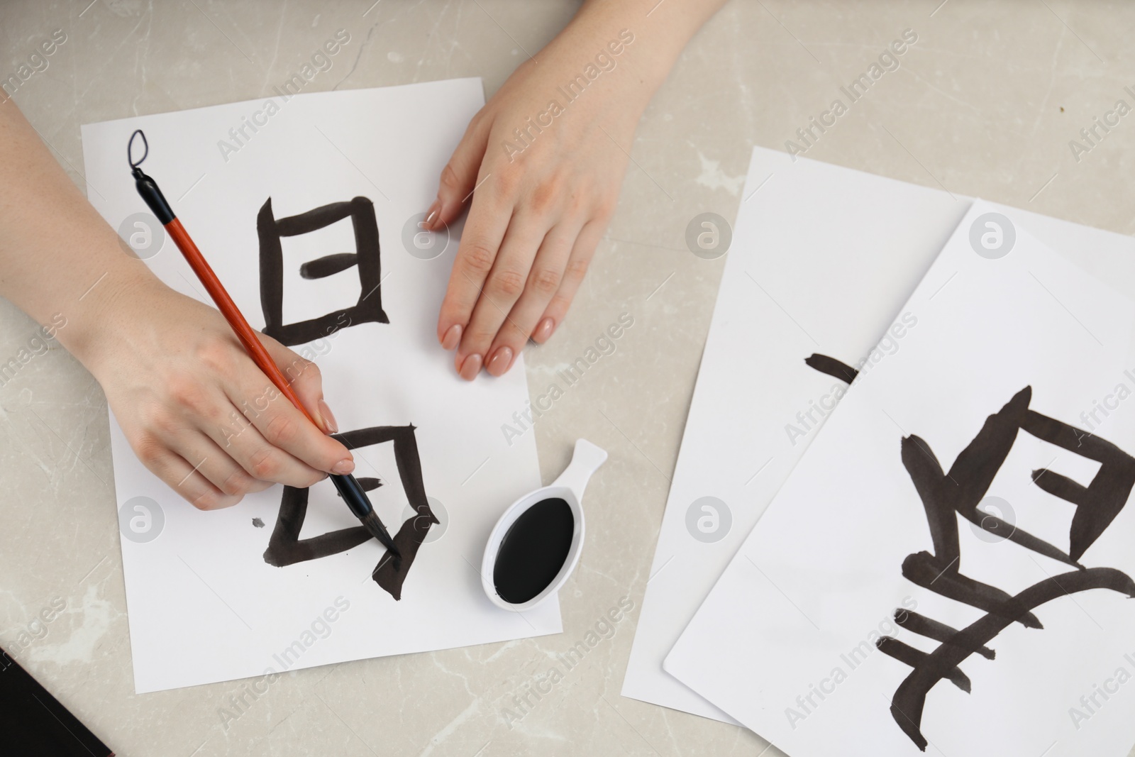 Photo of Calligraphy. Woman with brush and inkwell writing words Speak and Day in Chinese on paper at light marble table, top view