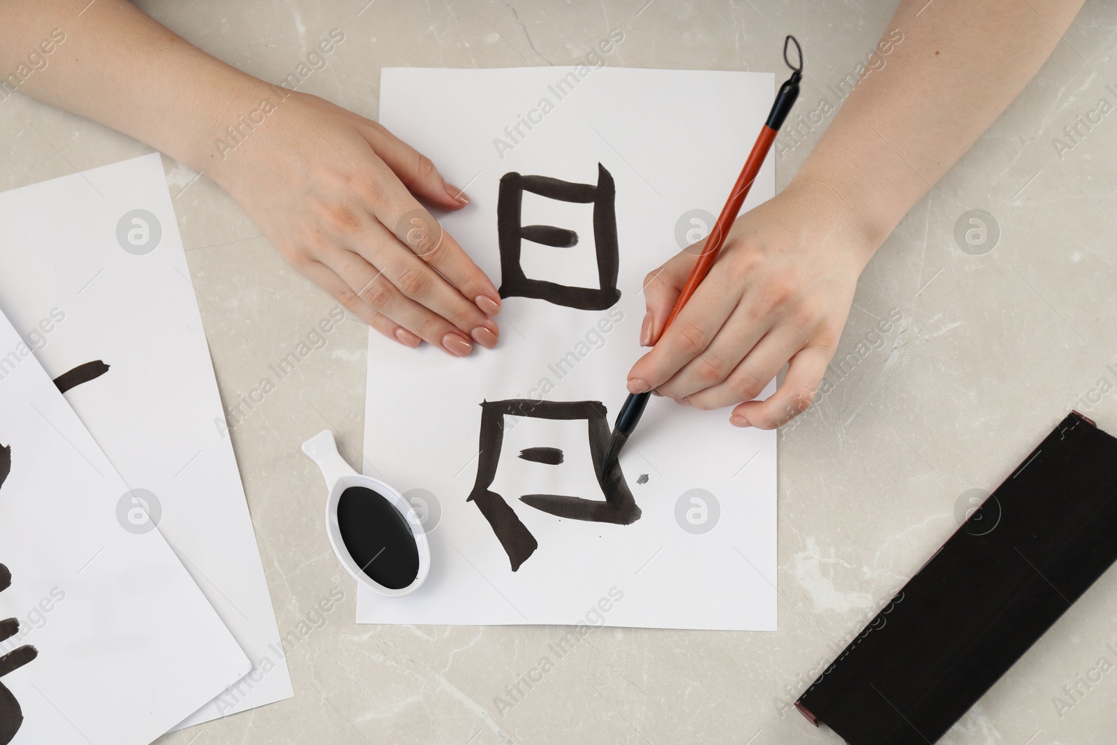 Photo of Calligraphy. Woman with brush and inkwell writing words Speak and Day in Chinese on paper at light marble table, top view