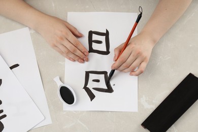 Calligraphy. Woman with brush and inkwell writing words Speak and Day in Chinese on paper at light marble table, top view