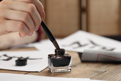 Photo of Calligraphy. Woman dipping brush into inkwell at wooden table, closeup