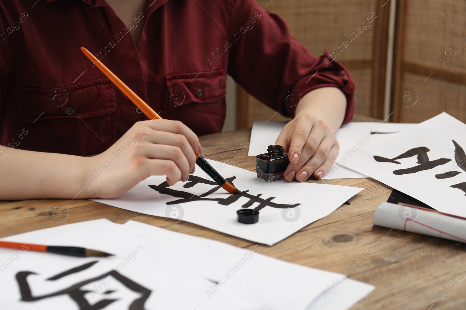 Photo of Calligraphy. Woman with brush and inkwell practicing writing Chinese hieroglyphs on paper at wooden table, closeup