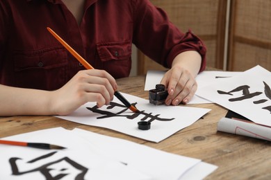 Calligraphy. Woman with brush and inkwell practicing writing Chinese hieroglyphs on paper at wooden table, closeup