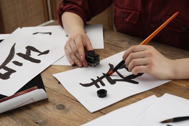 Photo of Calligraphy. Woman with brush and inkwell practicing writing Chinese hieroglyphs on paper at wooden table, closeup