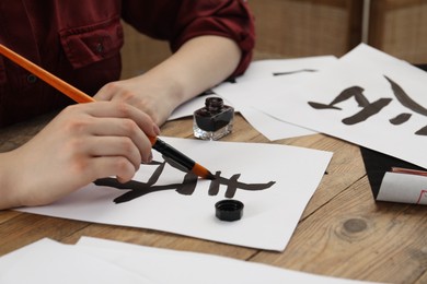 Calligraphy. Woman with brush and inkwell practicing writing Chinese hieroglyphs on paper at wooden table, closeup