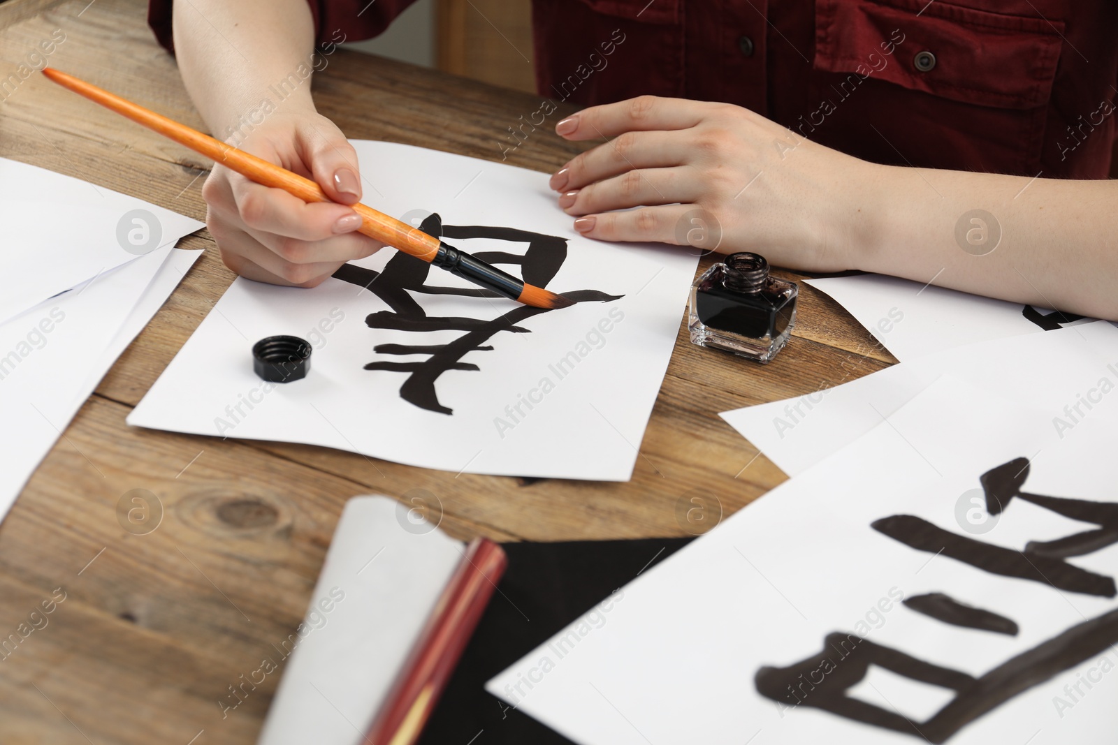 Photo of Calligraphy. Woman with brush and inkwell practicing writing Chinese hieroglyphs on paper at wooden table, closeup