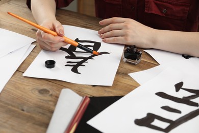 Calligraphy. Woman with brush and inkwell practicing writing Chinese hieroglyphs on paper at wooden table, closeup