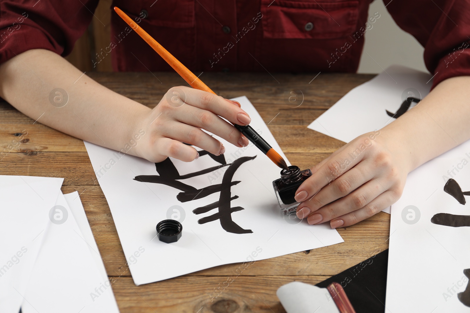 Photo of Calligraphy. Woman with brush and inkwell practicing writing Chinese hieroglyphs on paper at wooden table, closeup