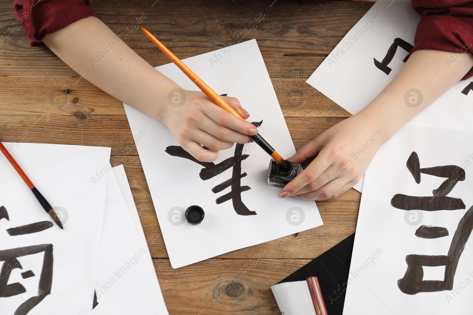 Photo of Calligraphy. Woman with brush and inkwell practicing writing Chinese hieroglyphs on paper at wooden table, top view