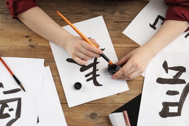 Calligraphy. Woman with brush and inkwell practicing writing Chinese hieroglyphs on paper at wooden table, top view