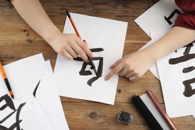 Photo of Calligraphy. Woman with brush and inkwell practicing writing Chinese hieroglyphs on paper at wooden table, top view