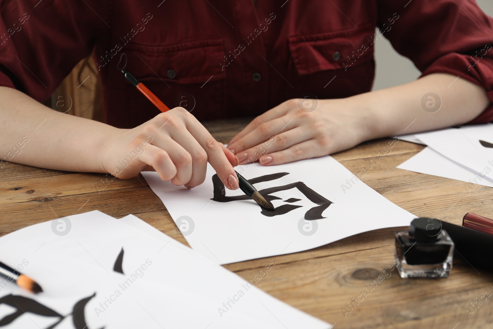 Photo of Calligraphy. Woman with brush and inkwell practicing writing Chinese hieroglyphs on paper at wooden table, closeup