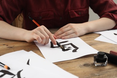 Calligraphy. Woman with brush and inkwell practicing writing Chinese hieroglyphs on paper at wooden table, closeup