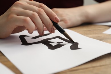 Photo of Calligraphy. Woman with brush writing word Spring in Chinese on paper at wooden table, closeup