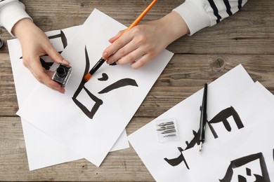 Photo of Calligraphy. Woman with brush and inkwell practicing writing Chinese hieroglyphs on paper at wooden table, top view