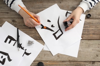 Photo of Calligraphy. Woman with brush and inkwell practicing writing Chinese hieroglyphs on paper at wooden table, top view