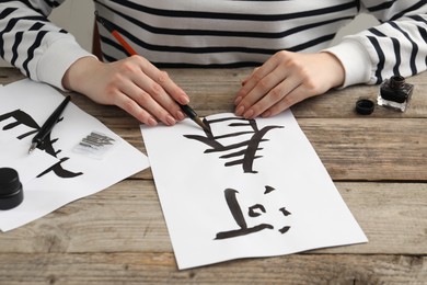 Calligraphy. Woman with brush and inkwell practicing writing Chinese hieroglyphs on paper at wooden table, closeup