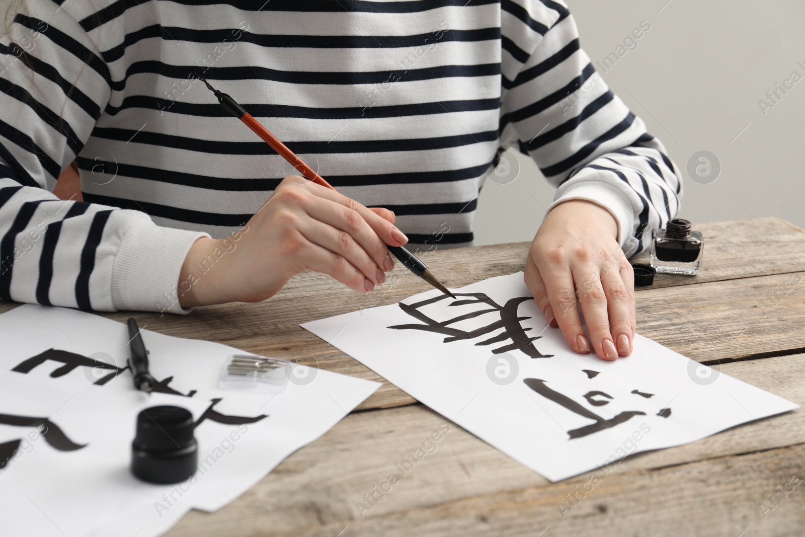 Photo of Calligraphy. Woman with brush and inkwell practicing writing Chinese hieroglyphs on paper at wooden table, closeup