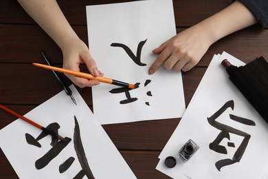 Photo of Calligraphy. Woman with brush and inkwell practicing writing Chinese hieroglyphs on paper at wooden table, top view