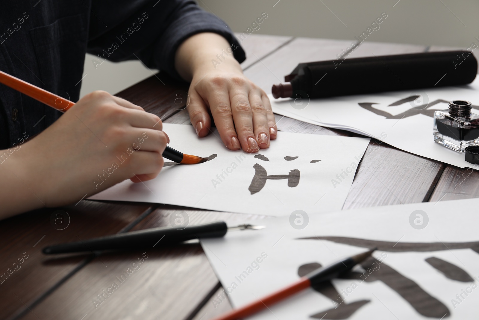 Photo of Calligraphy. Woman with brush and inkwell practicing writing Chinese hieroglyphs on paper at wooden table, closeup