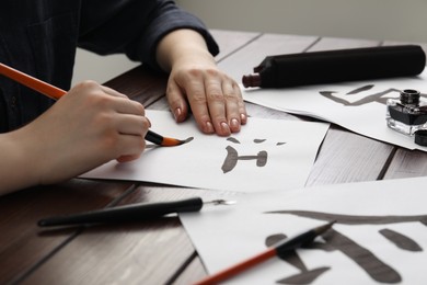 Photo of Calligraphy. Woman with brush and inkwell practicing writing Chinese hieroglyphs on paper at wooden table, closeup