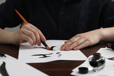 Calligraphy. Woman with brush and inkwell writing words River and Entrance in Chinese on paper at wooden table, closeup