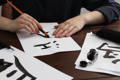 Photo of Calligraphy. Woman with brush and inkwell practicing writing Chinese hieroglyphs on paper at wooden table, closeup