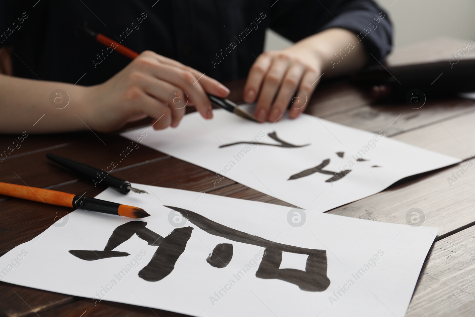 Photo of Calligraphy. Woman with brush practicing writing Chinese hieroglyphs on paper at wooden table, selective focus