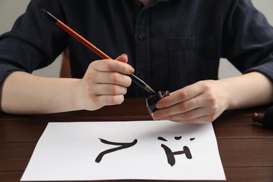 Calligraphy. Woman with brush and inkwell writing words River and Entrance in Chinese on paper at wooden table, closeup