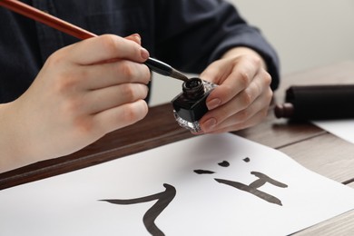 Calligraphy. Woman with brush and inkwell writing words River and Entrance in Chinese on paper at wooden table, closeup