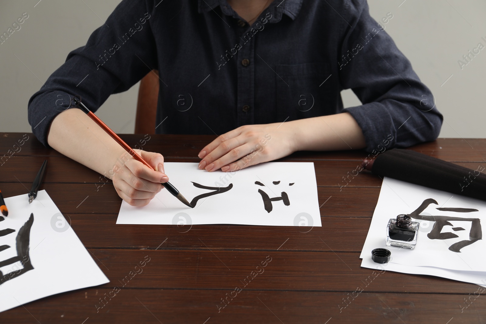 Photo of Calligraphy. Woman with brush and inkwell writing words River, Entrance and Bird in Chinese on paper at wooden table, closeup