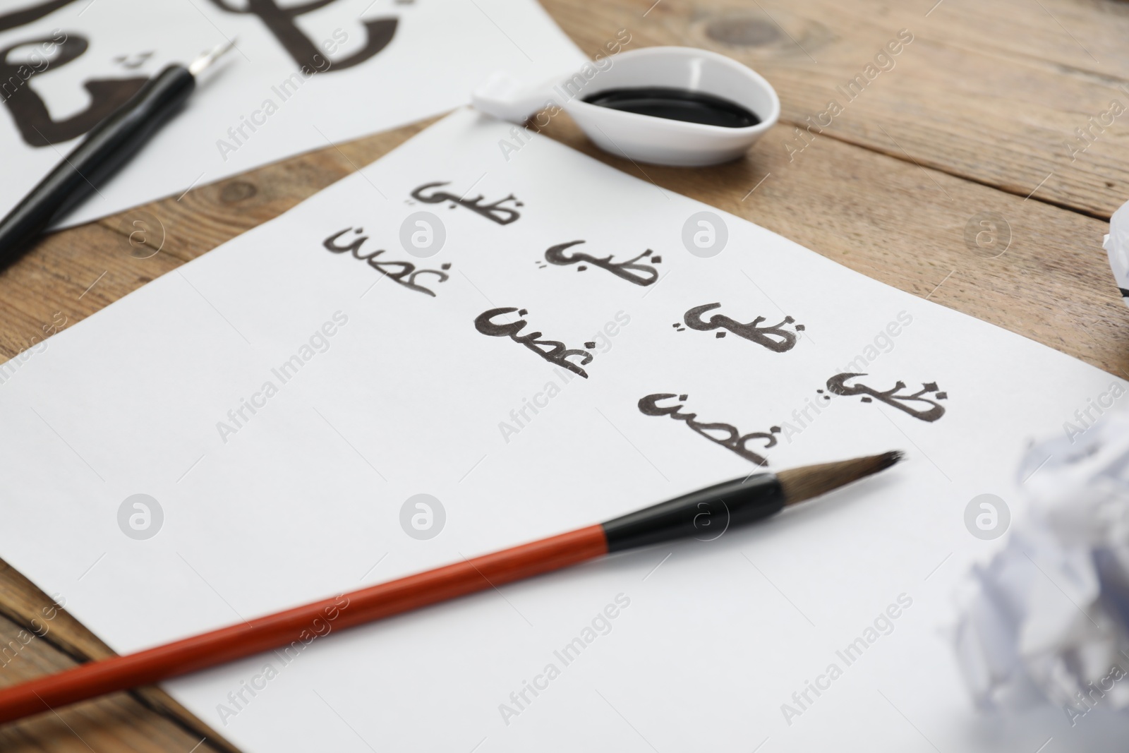 Photo of Calligraphy. Sheets of paper with written words Antilope and Branch in Arabian language, inkwell, fountain pen and brush on wooden table, closeup