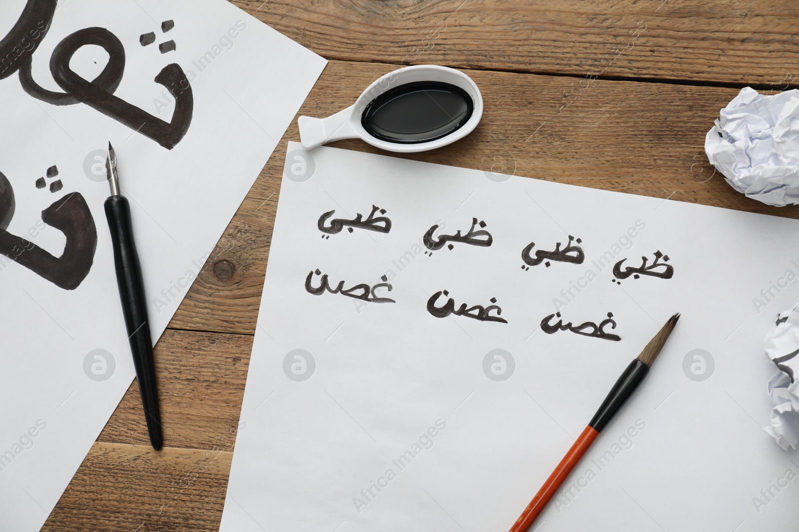 Photo of Calligraphy. Sheets of paper with written words Antilope and Branch in Arabian language, inkwell, fountain pen and brush on wooden table, flat lay