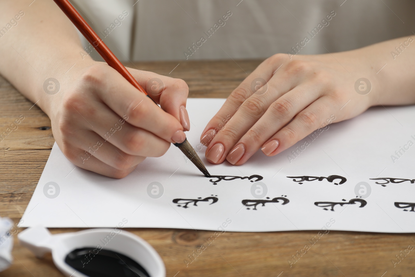 Photo of Calligraphy. Woman with brush writing words Antilope and Branch in Arabian language on paper at wooden table, closeup