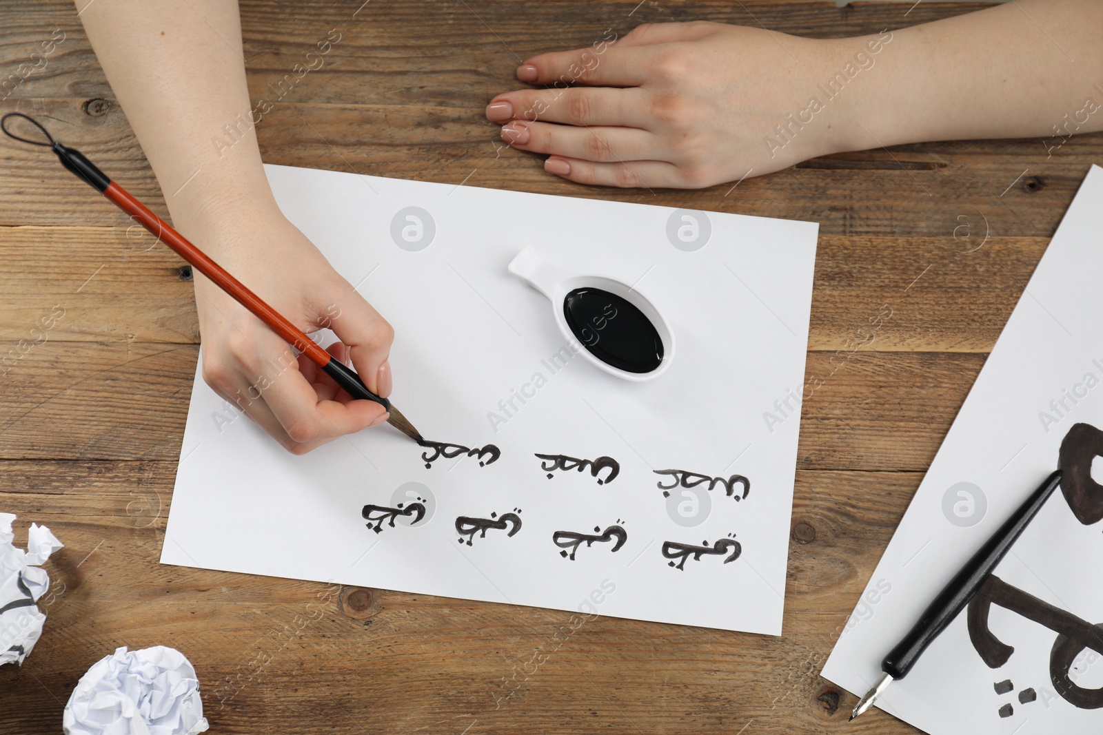 Photo of Calligraphy. Woman with brush writing words Antilope and Branch in Arabian language on paper at wooden table, top view