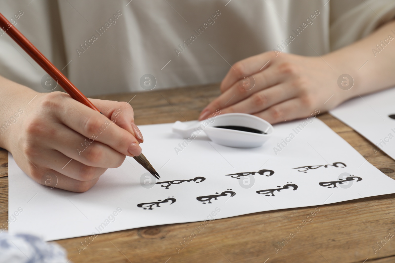 Photo of Calligraphy. Woman with brush writing words Antilope and Branch in Arabian language on paper at wooden table, closeup