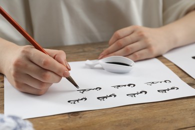 Calligraphy. Woman with brush writing words Antilope and Branch in Arabian language on paper at wooden table, closeup