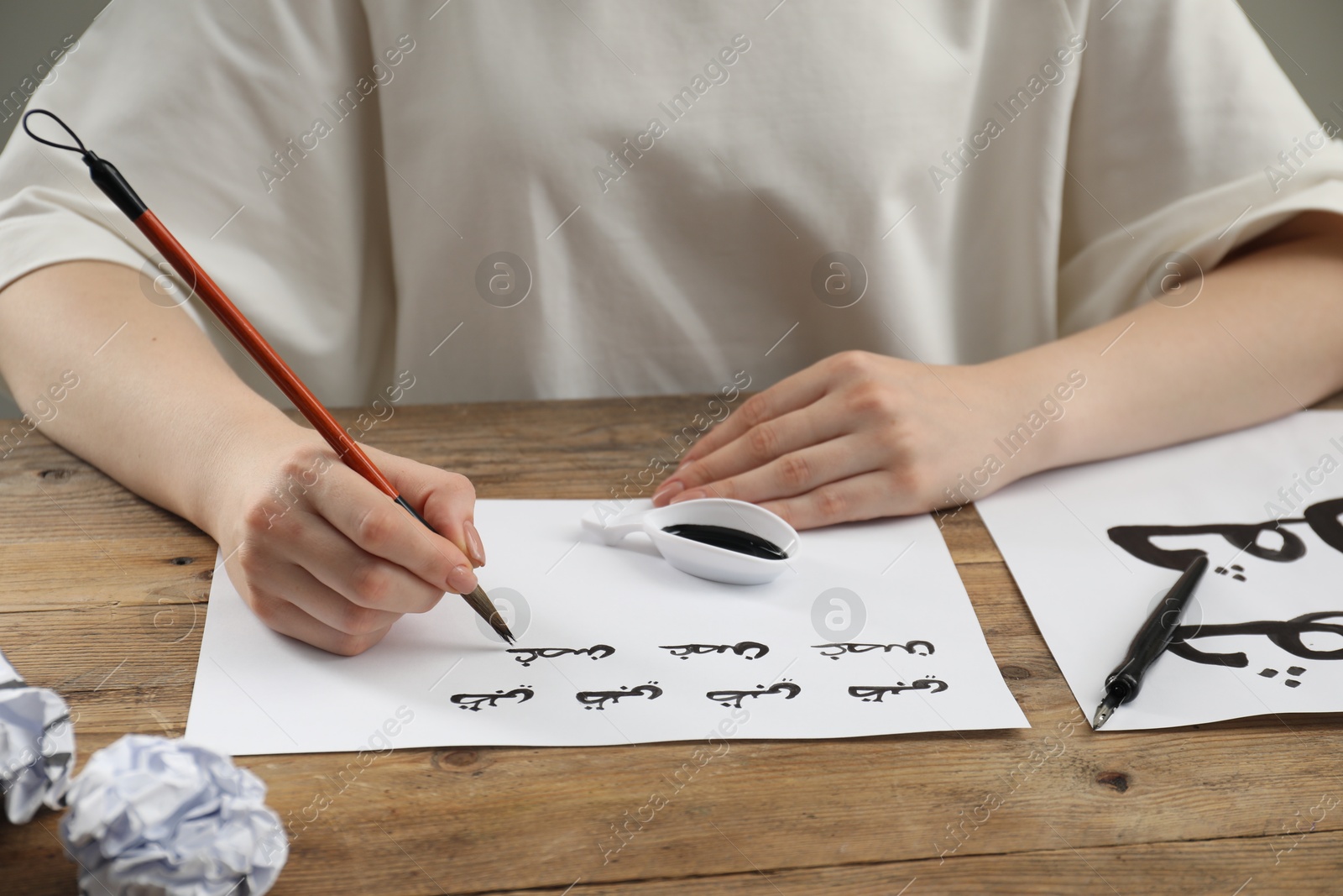 Photo of Calligraphy. Woman with brush writing words Antilope and Branch in Arabian language on paper at wooden table, closeup