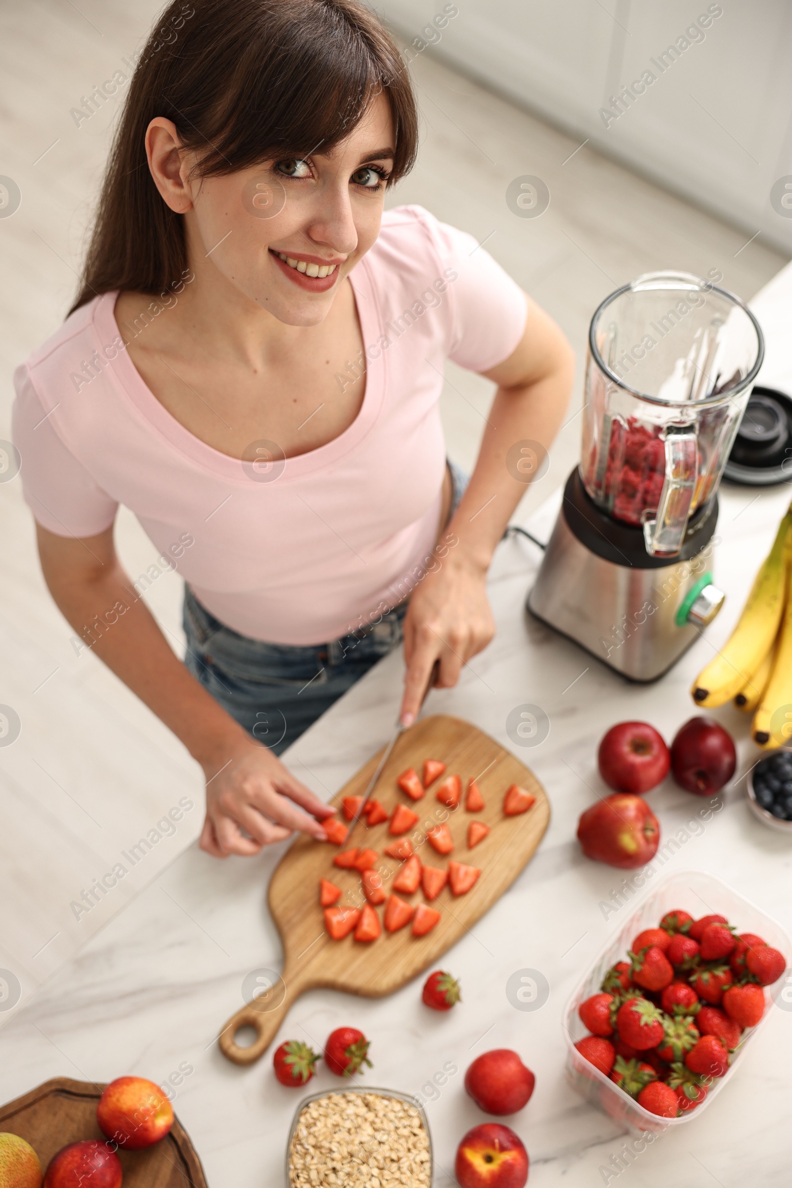 Photo of Young woman making delicious smoothie with blender at white marble table in kitchen, above view