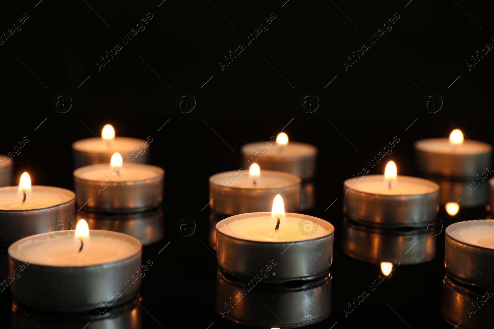 Photo of Many burning tealight candles on mirror surface against black background