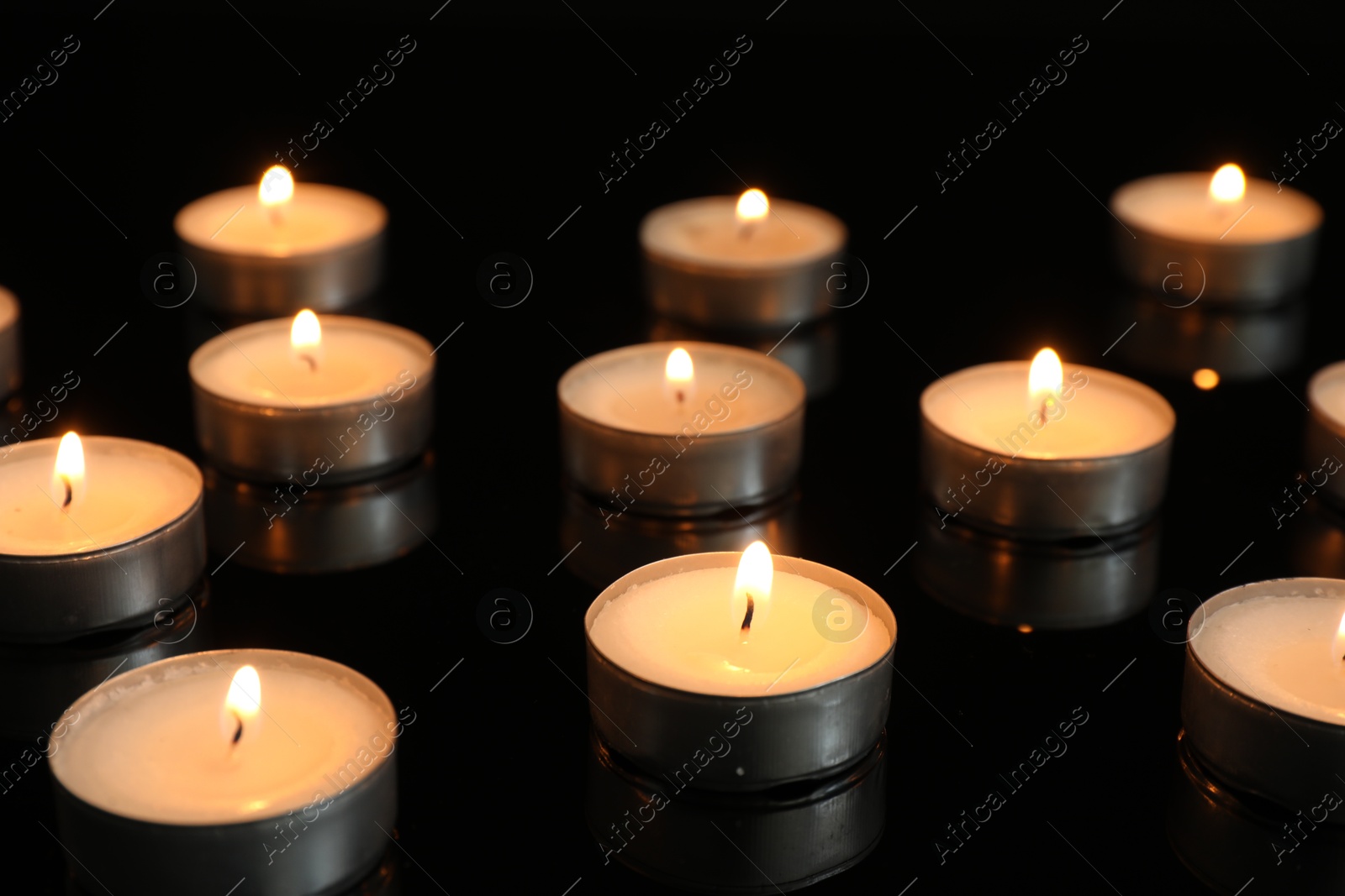 Photo of Many burning tealight candles on mirror surface against black background