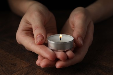 Photo of Woman holding burning tealight candle at wooden table, closeup