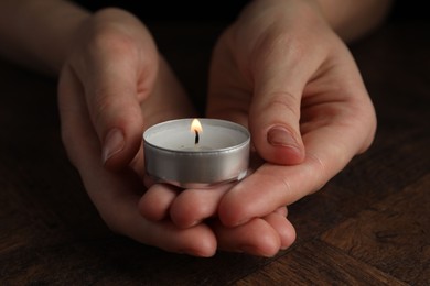 Photo of Woman holding burning tealight candle at wooden table, closeup