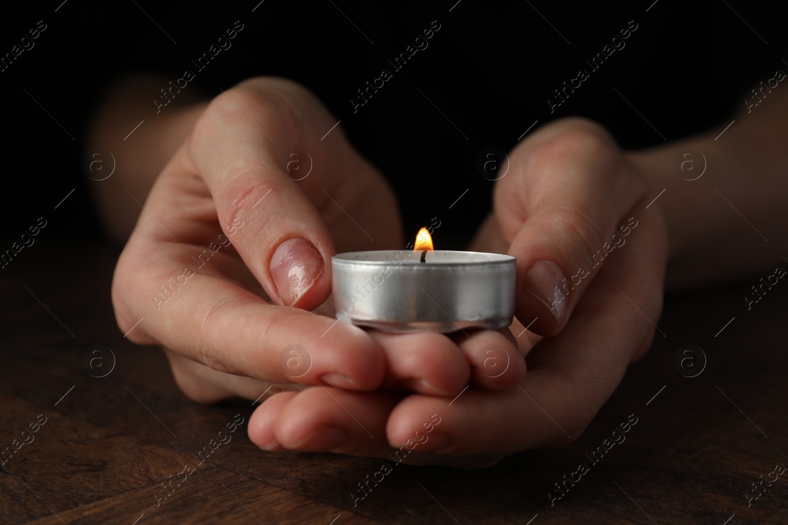 Photo of Woman holding burning tealight candle at wooden table, closeup