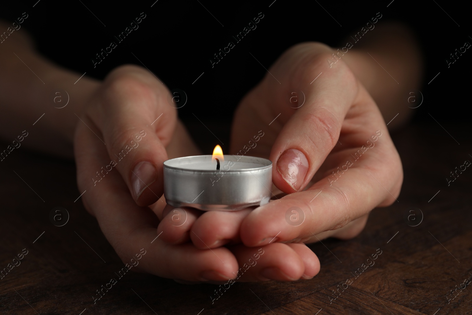 Photo of Woman holding burning tealight candle at wooden table, closeup